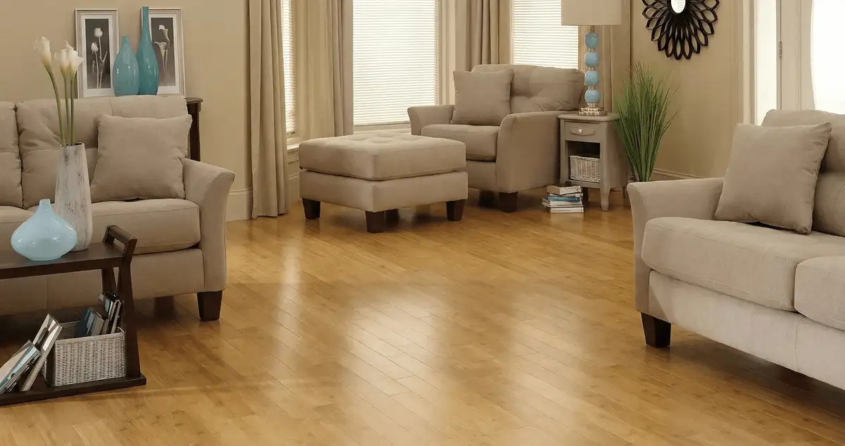 A living room with professionally installed bamboo flooring, beige sofas, a matching ottoman, a side table with a blue lamp, vases, framed pictures, and large windows with blinds and curtains in St Paul, MN